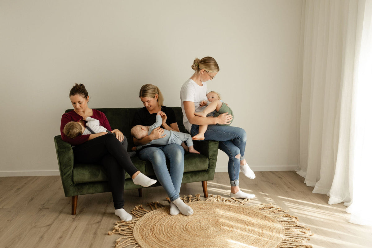 Three women sitting on a green sofa breastfeeding babies in a room with neutral decor.